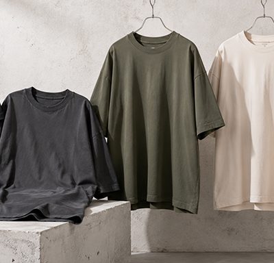 Five oversized t-shirts in black, charcoal, olive, beige, and white on hangers and a chair, displayed against a concrete wall as a product row.